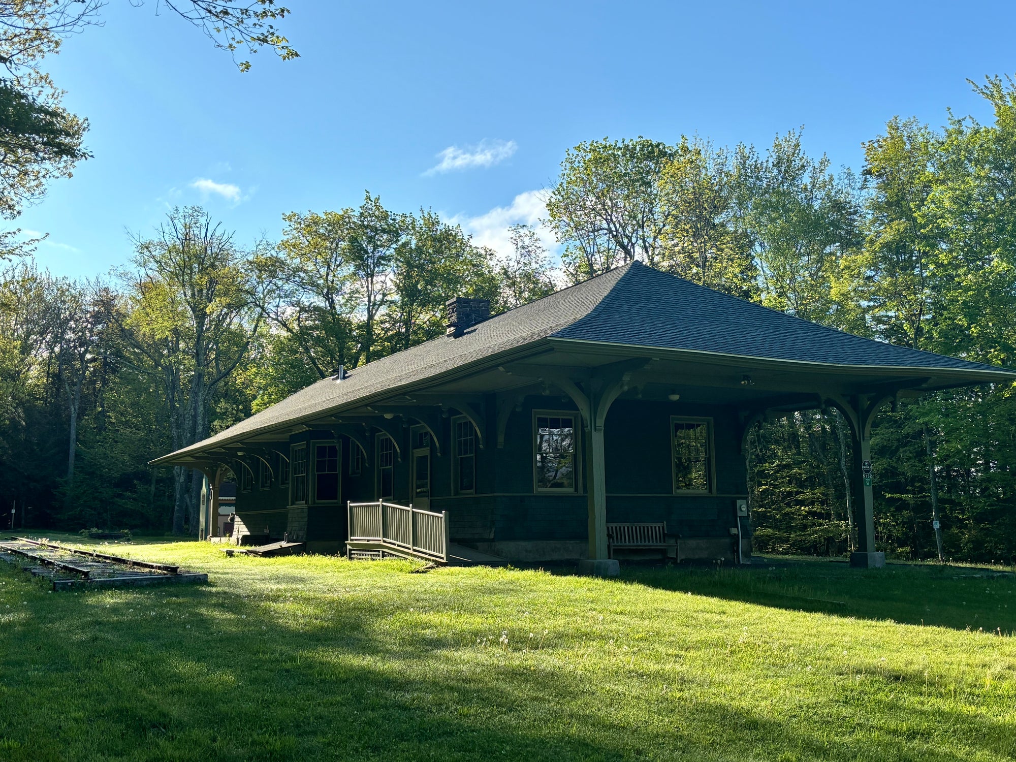 A single-story wooden train station with a wide covered porch, set on a grassy lawn with trees and blue sky in the background.