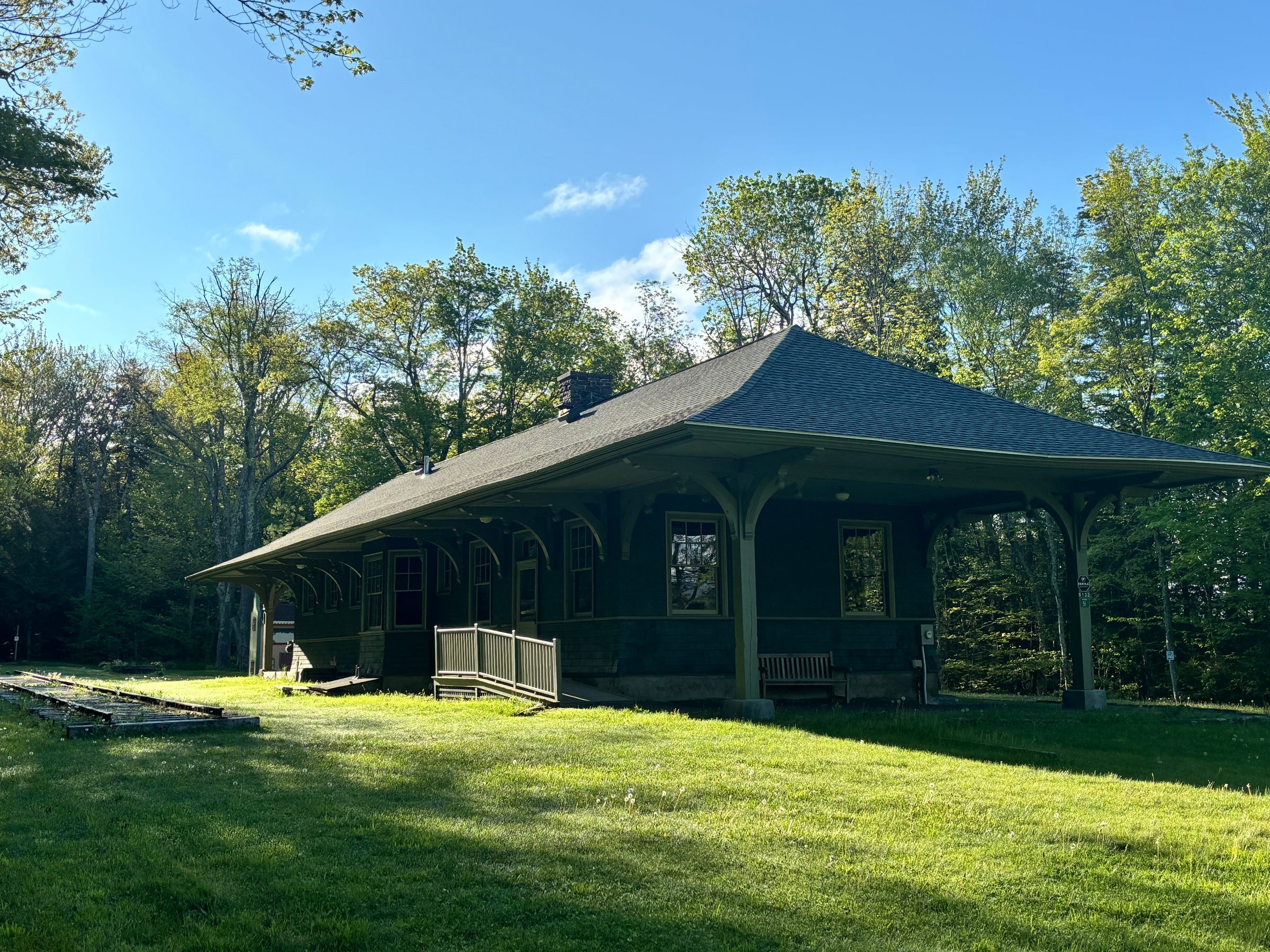 A single-story wooden train station with a wide covered porch, set on a grassy lawn with trees and blue sky in the background.