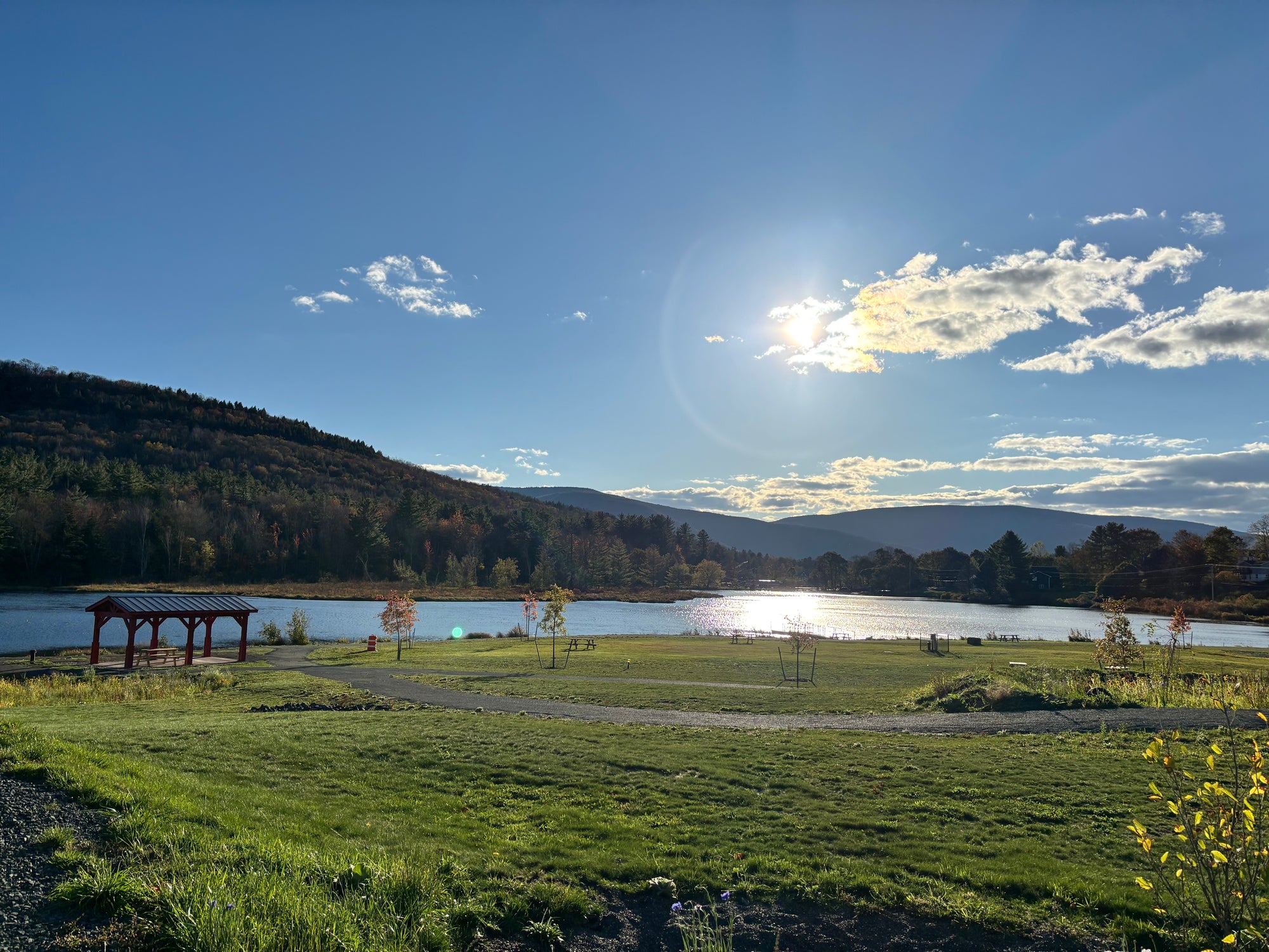 A scenic outdoor landscape with a grassy field, a small wooden pavilion, a lake, forested hills, and a bright sun in a blue sky with scattered clouds.
